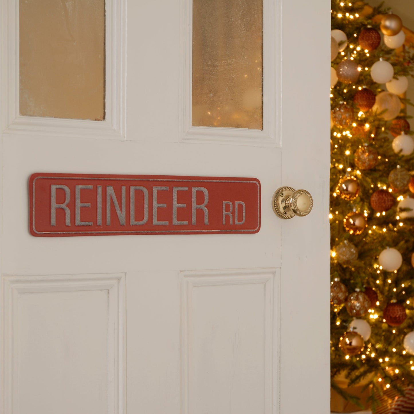 A red metal Christmas sign with 'Reindeer Rd' written on it, mounted on a door with a Christmas tree in the background.