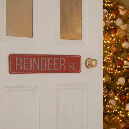 A red metal Christmas sign with 'Reindeer Rd' written on it, mounted on a door with a Christmas tree in the background.