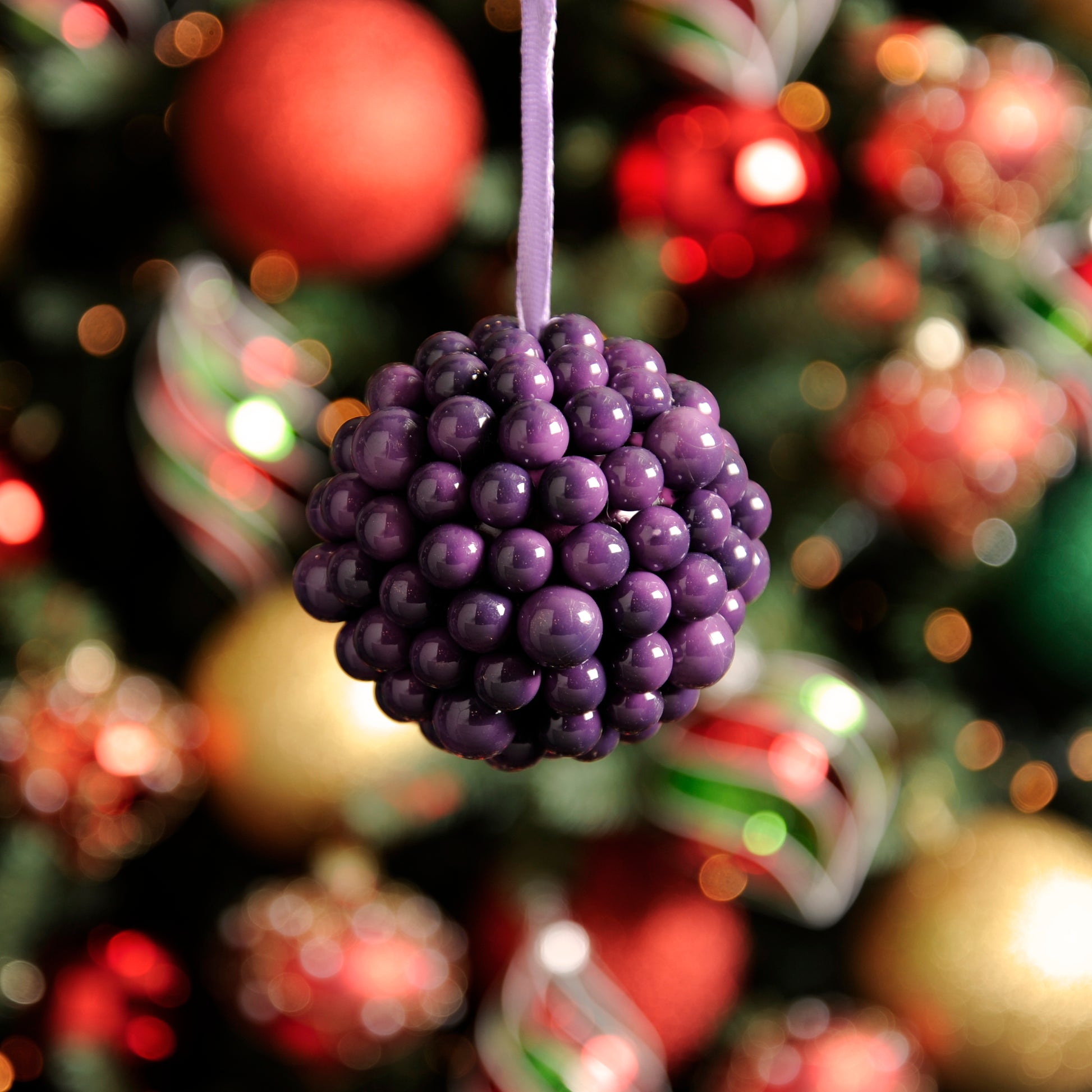 A purple berry cluster ball hanging from a Christmas tree, with a blurred background of multicolored ornaments.