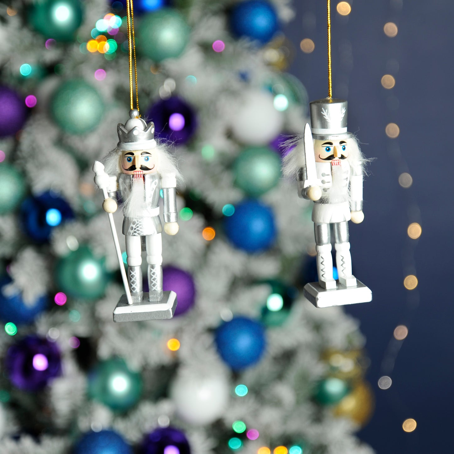 A pair of white and silver nutcrackers hanging from a Christmas tree, dressed in uniforms with swords and staff.