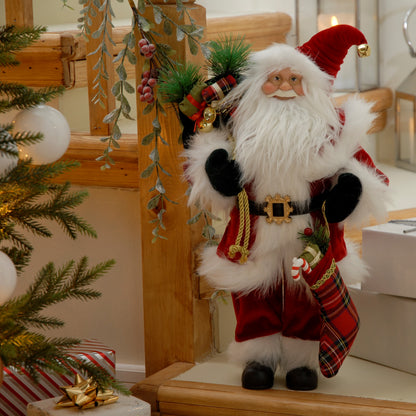 A traditional Santa figurine standing by a Christmas tree, wearing a red suit with a black belt, white hair, and a tartan stocking. The figure is holding a sack of presents and the stocking.