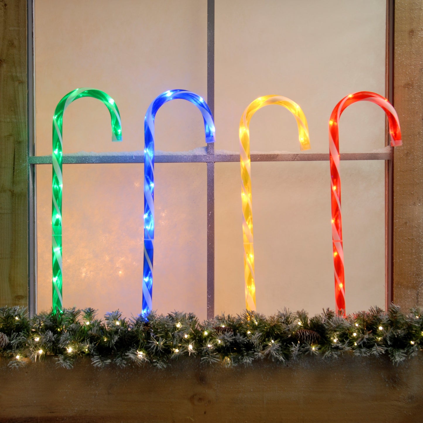 A set of four multi-colored LED candy cane stake lights displayed outdoors, with colors green, blue, red, and yellow, set against a backdrop of a Christmas decoration.