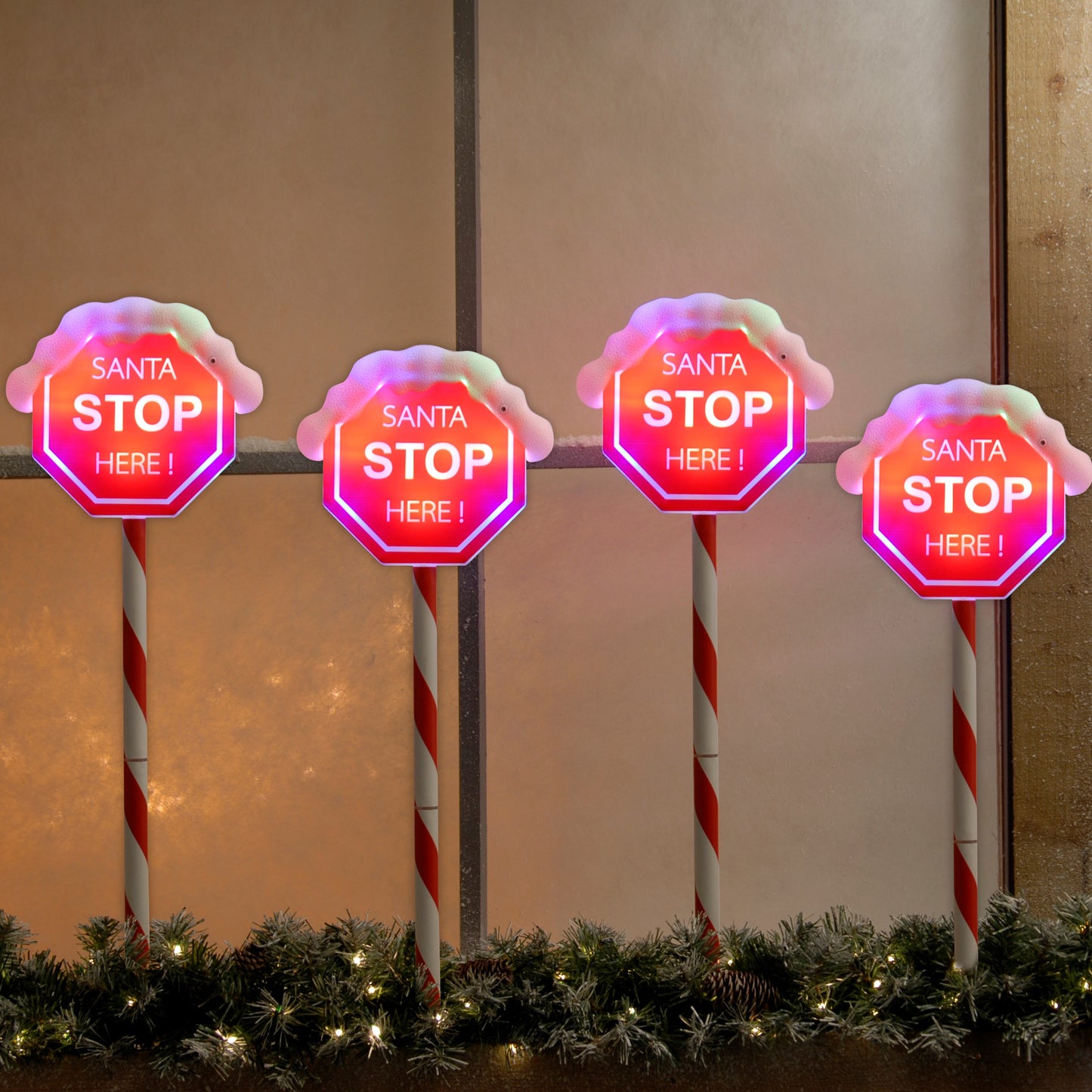 A set of four LED lit path stake lights with a Santa Stop Here message, featuring red and white candy stripes on poles with a simulated snowy ground and Christmas decorations in the background.