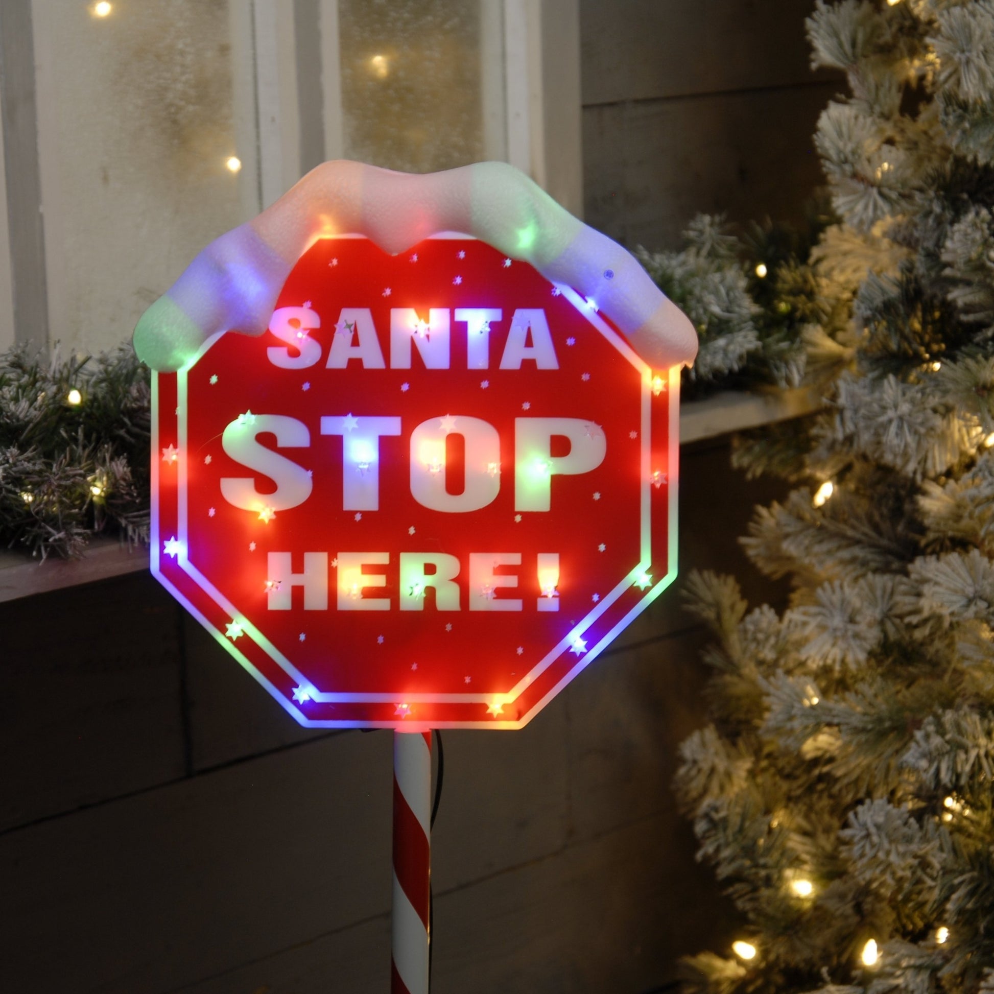 A lit LED sign with the text 'Santa Stop Here!' in red and white, mounted on a candy cane pole, with multicolor lights and artificial snow on top.