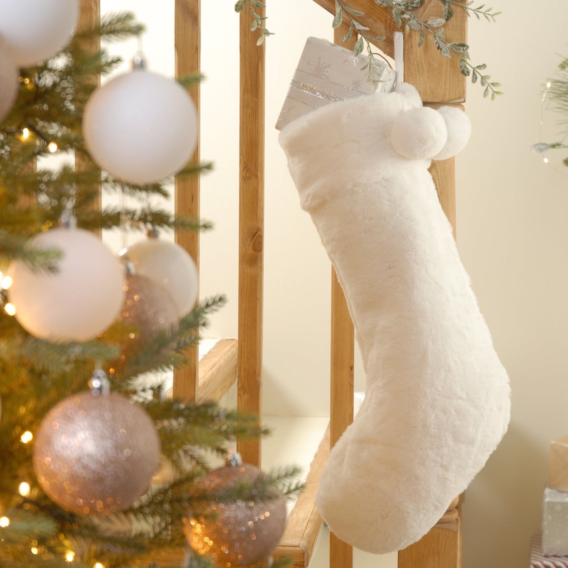 A white faux fur Christmas stocking hanging from a fireplace, with decorative pompoms and a tree skirt in the background.