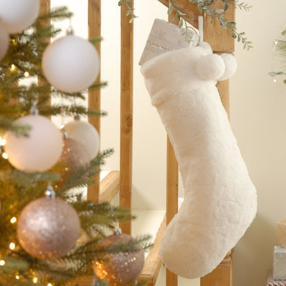 A white faux fur Christmas stocking hanging from a fireplace, with decorative pompoms and a tree skirt in the background.