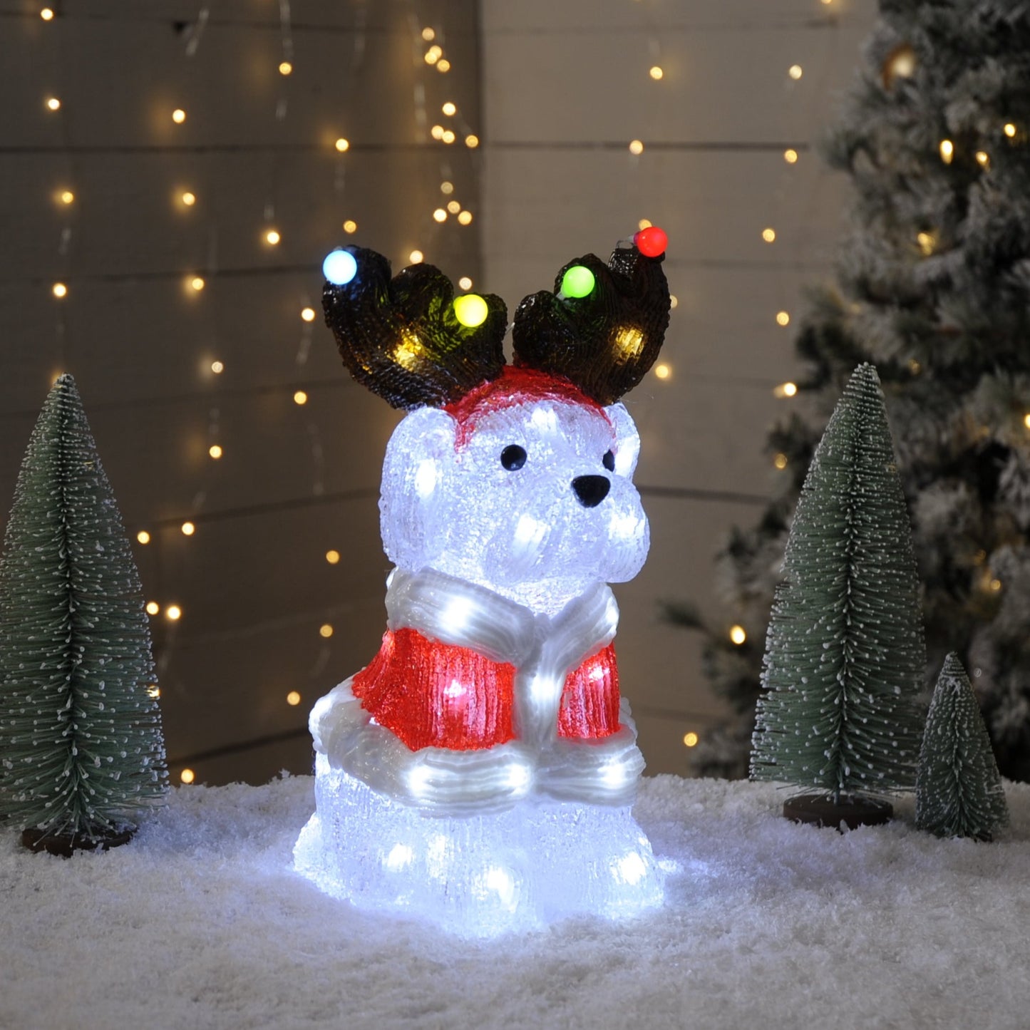 A lit acrylic sitting dog decoration with multicolored LED lights, displayed on a snowy surface with Christmas trees in the background.