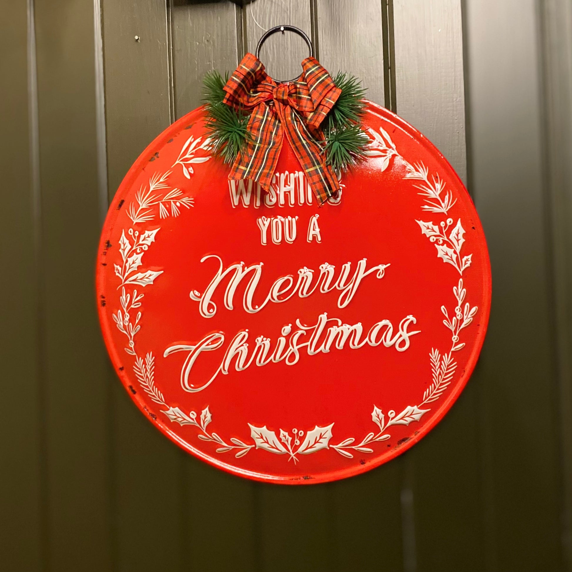 A round, red metal Christmas sign with white text reading 'Wishing you a Merry Christmas' decorated with festive bows and seasonal winter foliage.