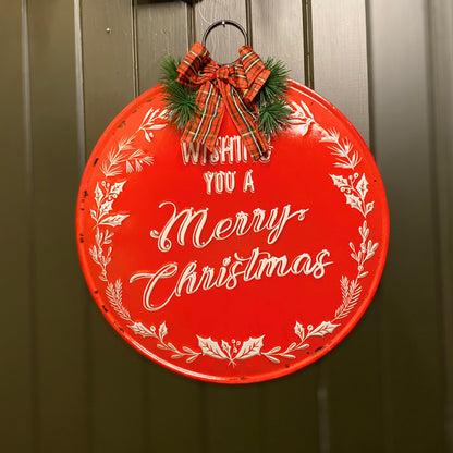 A round, red metal Christmas sign with white text reading 'Wishing you a Merry Christmas' decorated with festive bows and seasonal winter foliage.