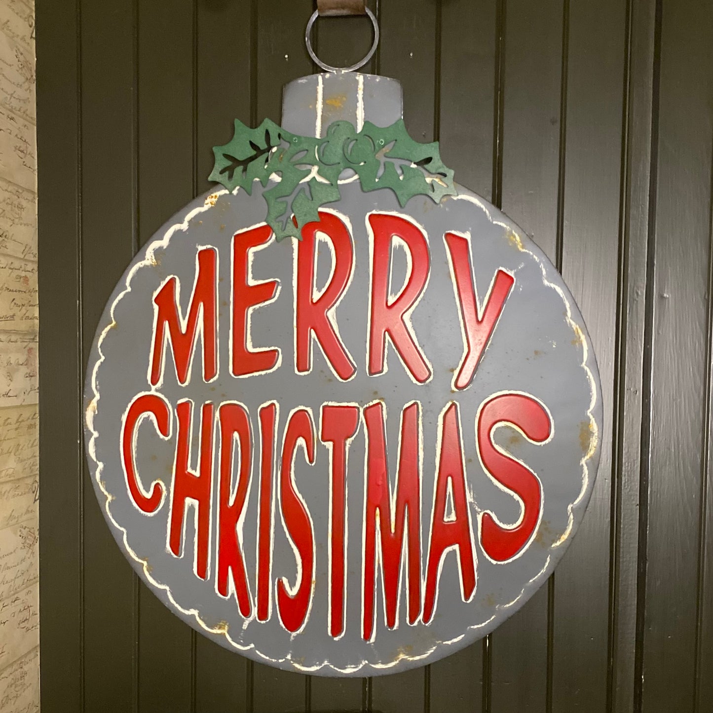 A round, rustic metal Christmas sign with 'Merry Christmas' text in red, aged patina, and a holly bow at the top.