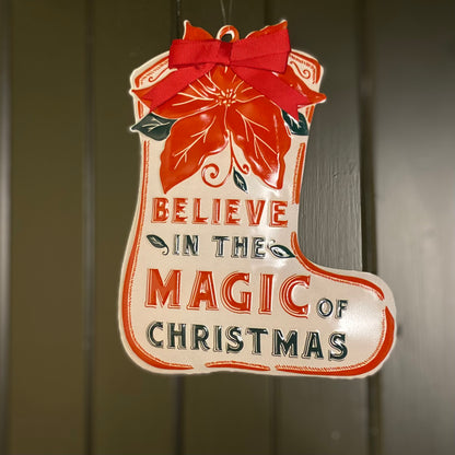 A red, white, and green metal sign with the text 'BELIEVE IN THE MAGIC OF CHRISTMAS' and a floral design.