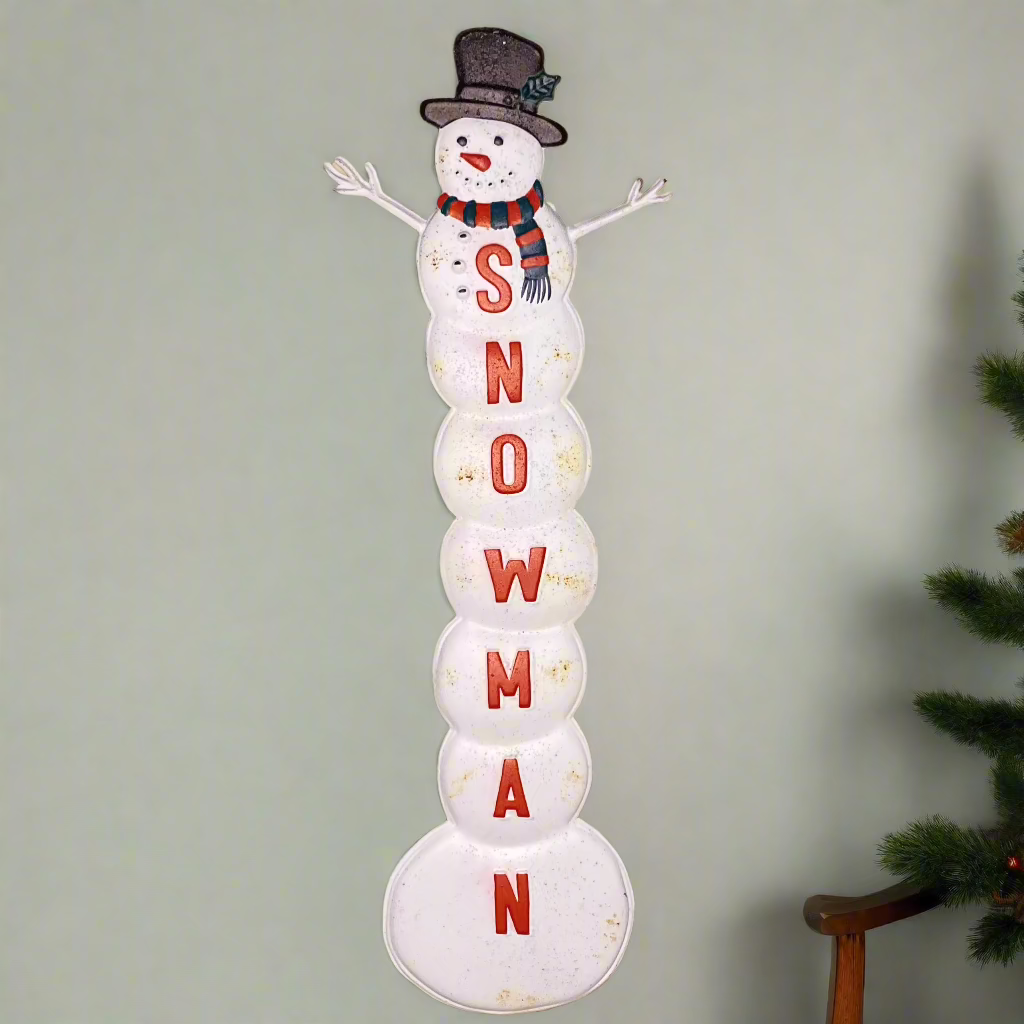 A vintage-style white snowman Christmas sign with 'Snowman' text in red, placed against a neutral background with a Christmas tree in the corner.