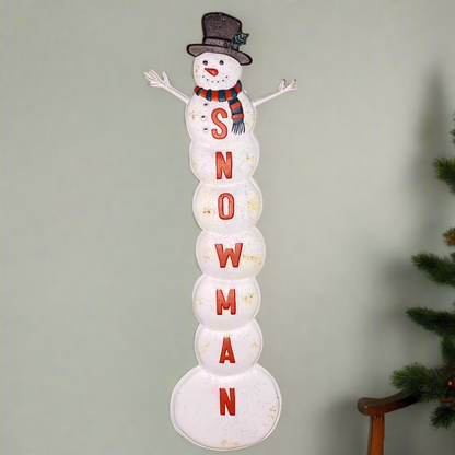 A vintage-style white snowman Christmas sign with 'Snowman' text in red, placed against a neutral background with a Christmas tree in the corner.