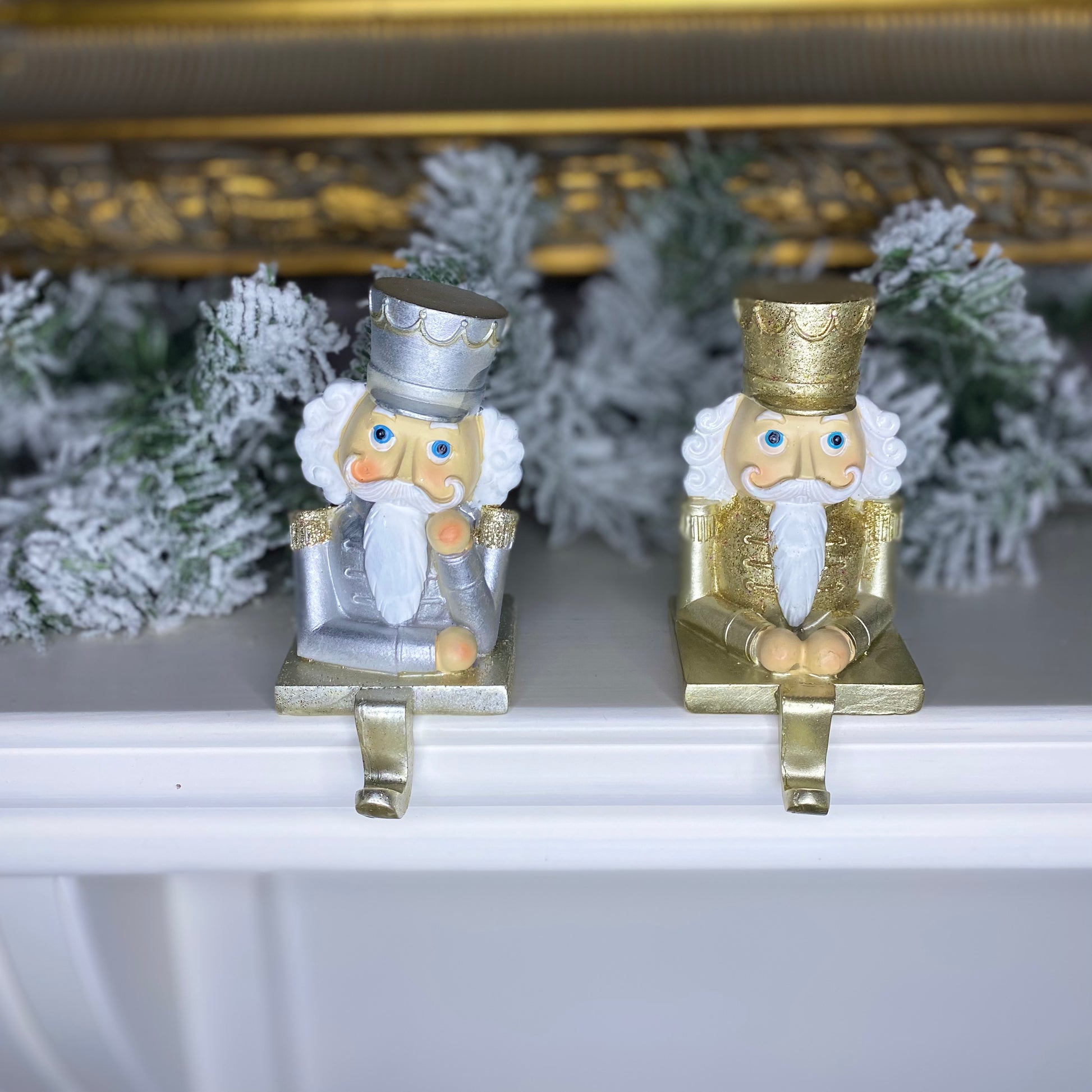 A set of two nutcracker stocking hangers, one in gold and one in silver, placed on a mantelpiece with a decorative backdrop.