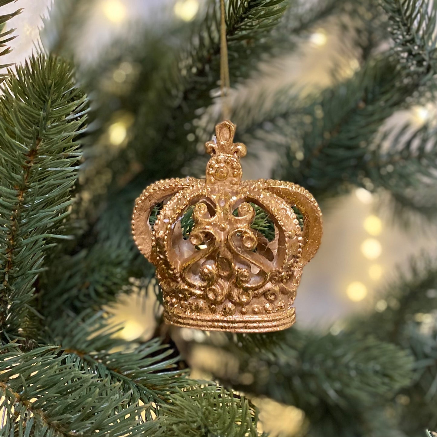 A golden hanging Christmas tree decoration in the shape of a crown, set against a blurred background of Christmas lights.