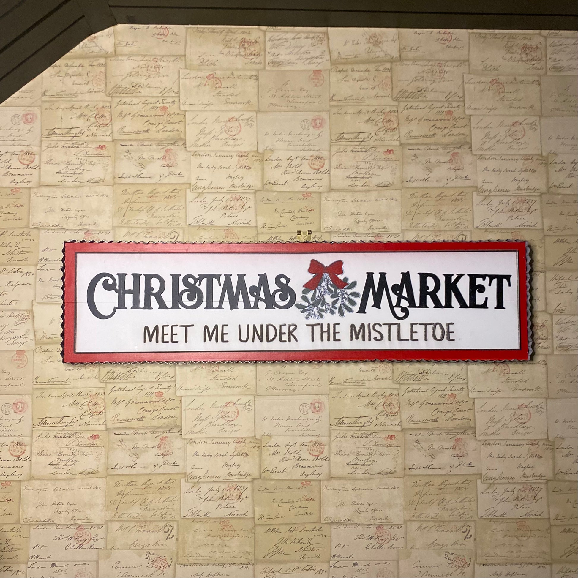 A white wooden sign with red and silver text and accents, hanging on a wall, promoting a Christmas market.