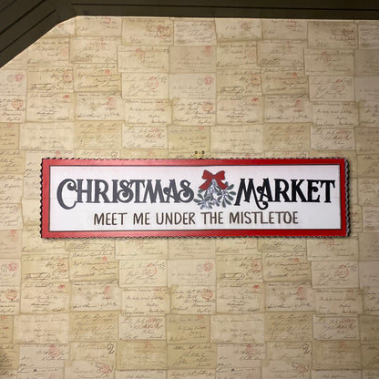 A white wooden sign with red and silver text and accents, hanging on a wall, promoting a Christmas market.