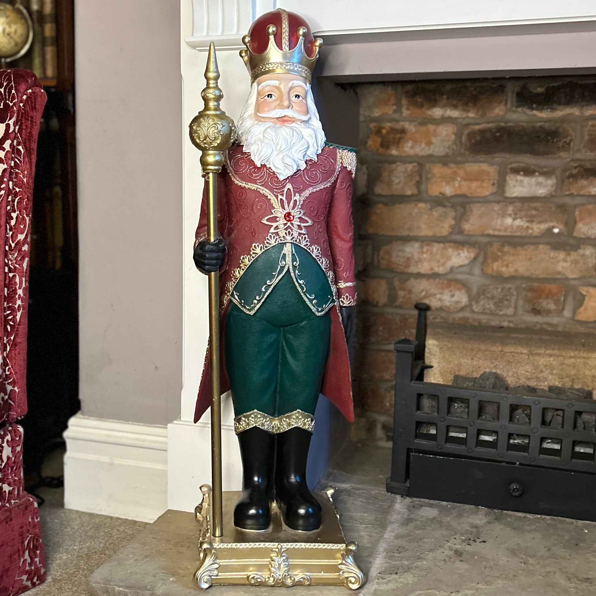 A decorative resin nutcracker figurine dressed in a colorful jacket with tassels, green velvet trousers, a majestic staff, and wearing a royal crown, standing on a golden plinth.