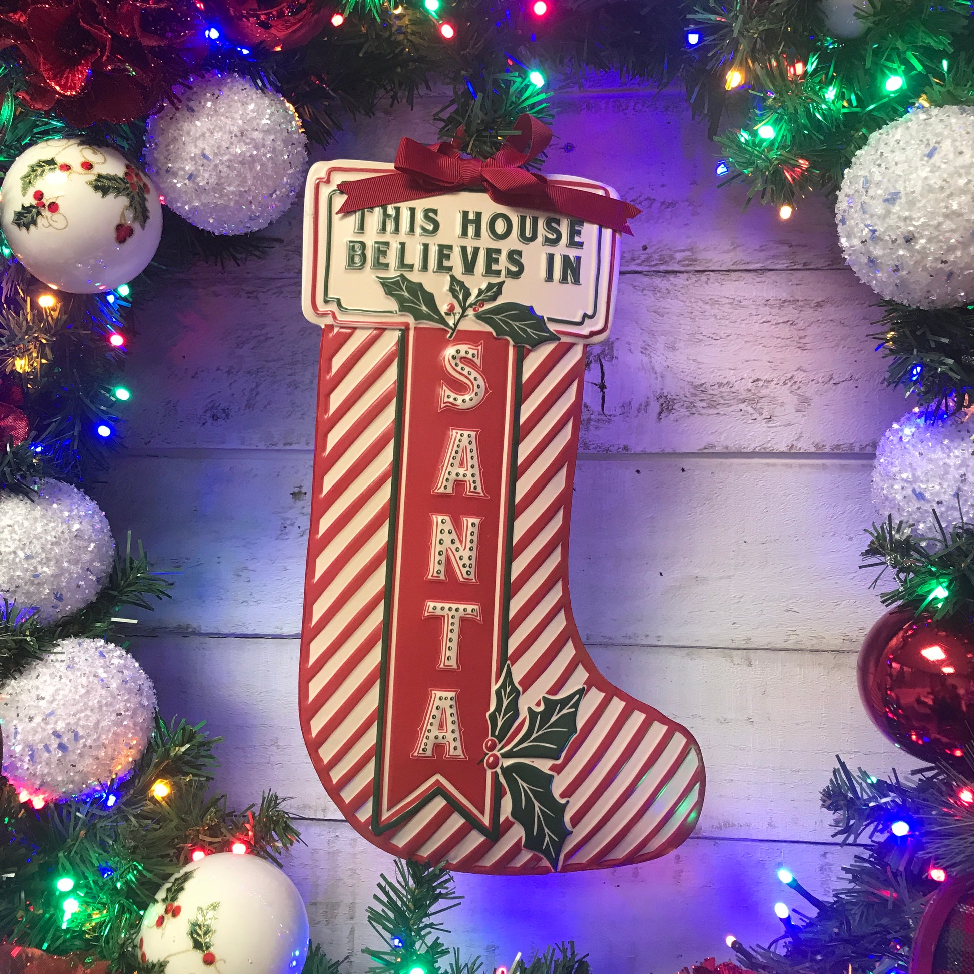 A red and white Christmas stocking shape sign with the text 'This house believes in Santa' embossed on it, displayed against a festive background with holiday ornaments.