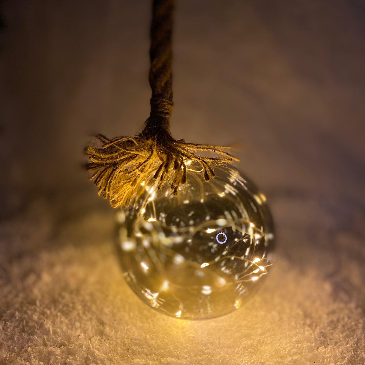 An illuminated LED ball Christmas decoration with a jute rope, emitting a warm white glow, placed on a white surface in a dark environment.