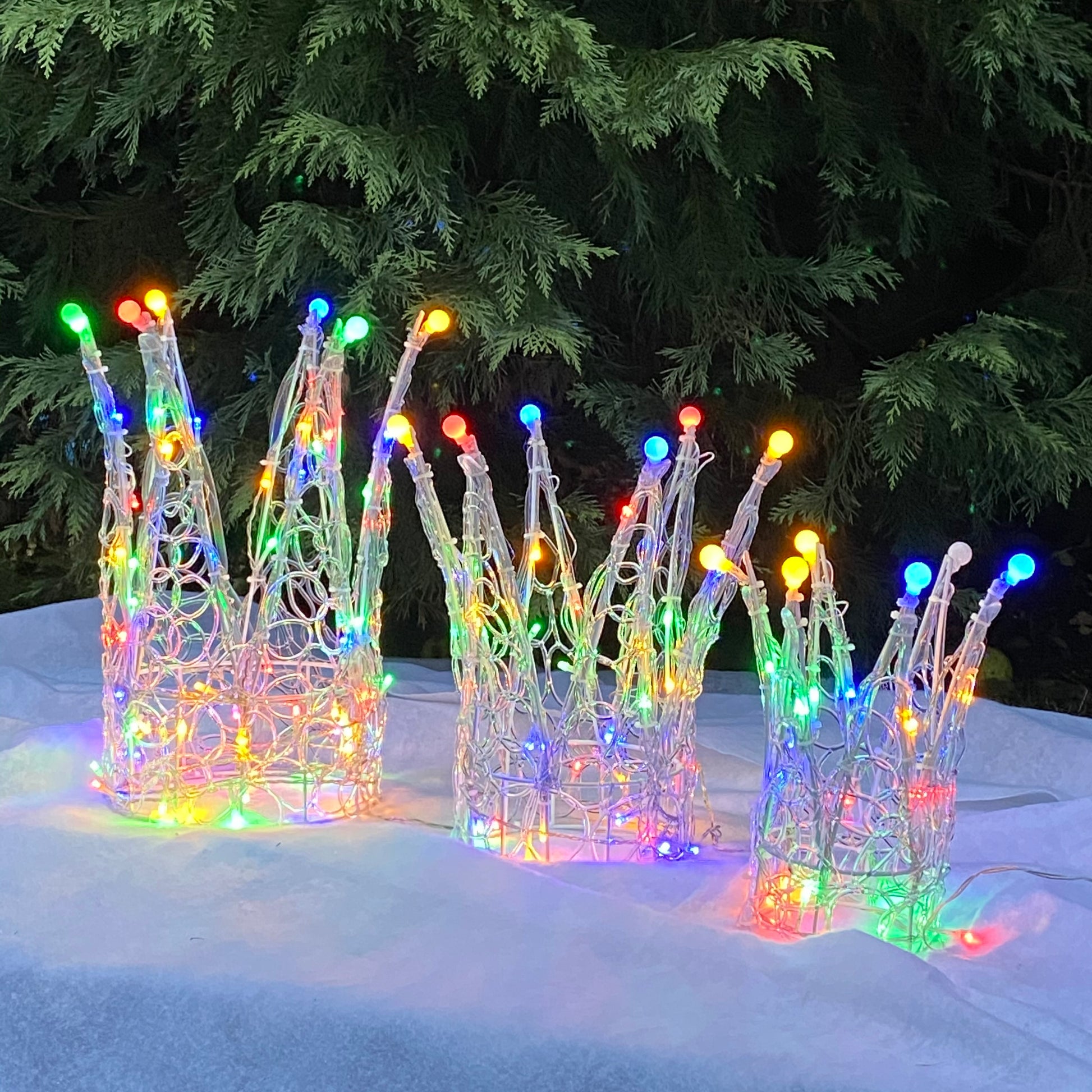 A set of three Christmas-themed crowns made from soft acrylic, each adorned with multi-colored LED lights, displayed outdoors against a backdrop of green pine trees.