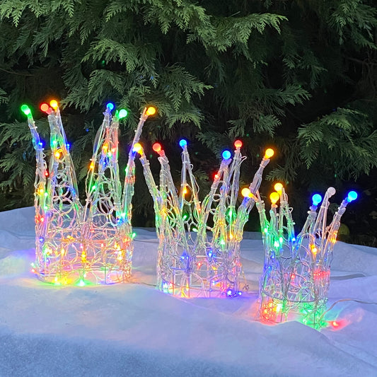 A set of three Christmas-themed crowns made from soft acrylic, each adorned with multi-colored LED lights, displayed outdoors against a backdrop of green pine trees.