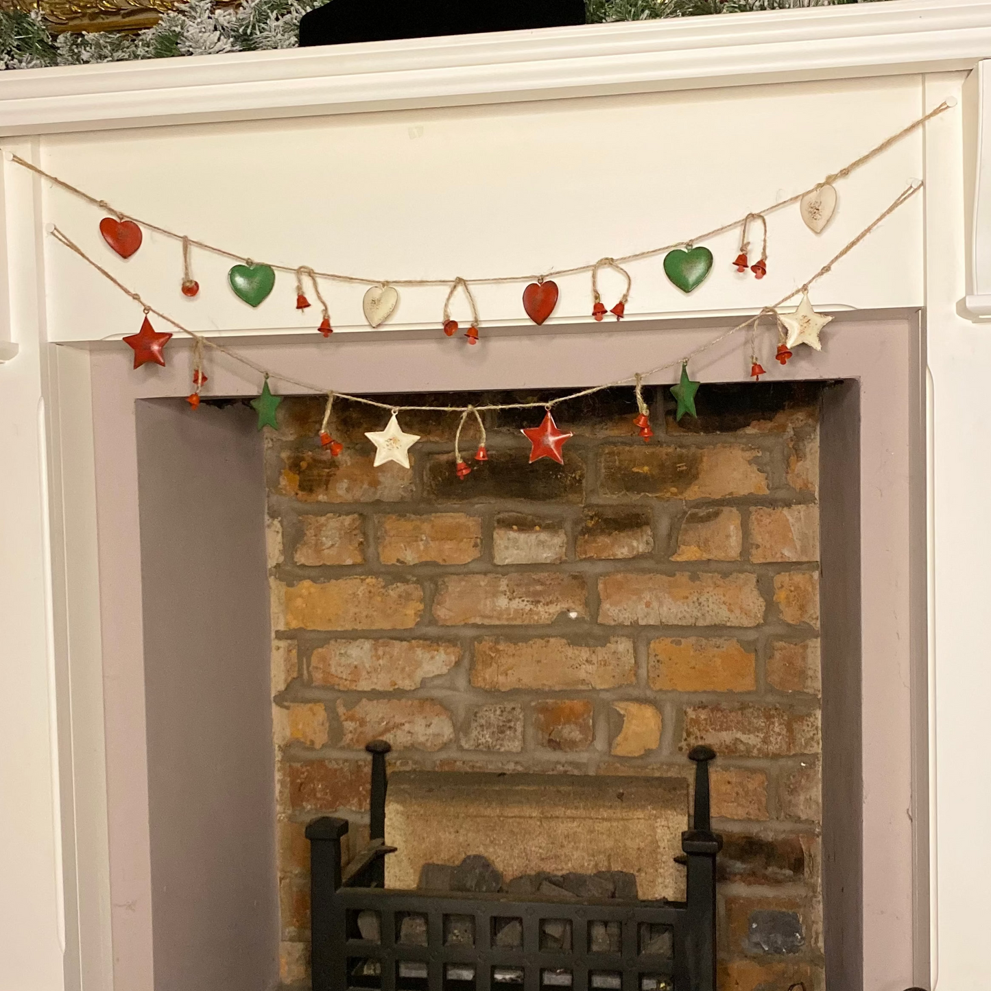 A garland of red, white, and green hearts and stars hanging over a fireplace, with Christmas tree decorations in the background.